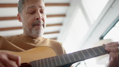 Low-angle-of-focused-senior-biracial-man-sitting-playing-guitar-at-home,-slow-motion