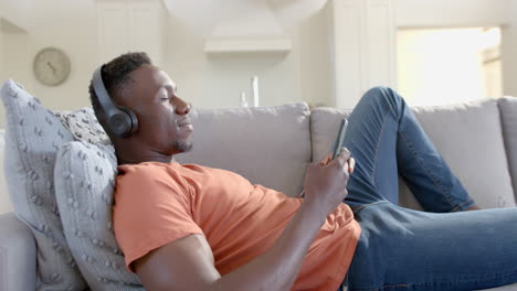 Happy-african-american-man-using-smartphone-and-headphones-in-sunny-living-room,-slow-motion