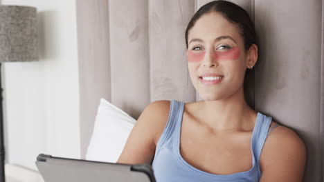 Portrait-of-happy-biracial-woman-with-under-eye-patches-sitting-on-bed-using-tablet,-slow-motion