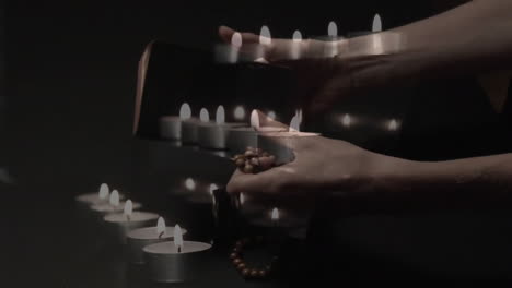 Animation-Brennender-Kerzen-über-Händen,-Die-Bibel-Und-Rosenkranz-Auf-Schwarzem-Hintergrund-Halten