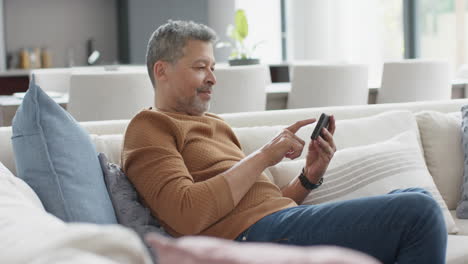 Glücklicher-älterer-Mann-Mit-Gemischter-Abstammung,-Der-Sein-Smartphone-Auf-Der-Couch-Im-Sonnigen-Wohnzimmer-Benutzt,-Zeitlupe
