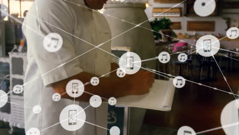 Animation-of-network-of-connections-with-icons-over-caucasian-cook-in-kitchen