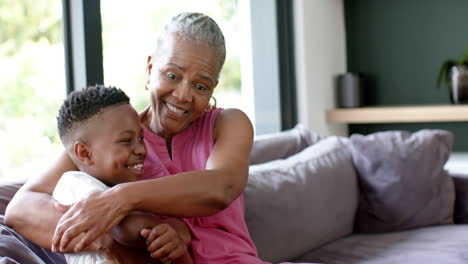Happy-african-american-grandson-and-grandmother-cuddling-on-sofa,-copy-space,-slow-motion