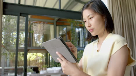 Happy-asian-woman-standing-and-using-tablet-in-sunny-living-room,-slow-motion
