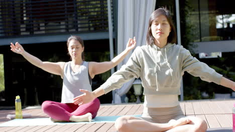 Amigas-Asiáticas-Enfocadas-Practicando-Meditación-De-Yoga-En-Una-Terraza-Soleada,-Cámara-Lenta