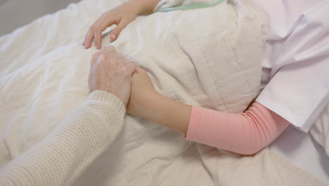 Hand-of-caucasian-mother-holding-hand-of-daughter-patient-lying-in-hospital-bed,-slow-motion