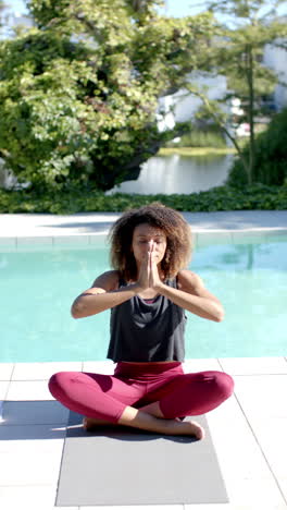 Vertikales-Video-Einer-Biracial-Frau,-Die-Yoga-Meditation-Praktiziert-Und-Am-Sonnigen-Pool-Sitzt,-Zeitlupe