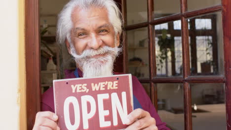 Happy-senior-biracial-business-owner-with-long-beard-holding-shop-sign-and-smiling-slow-motion