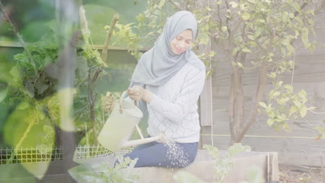 Animation-of-leaves-over-biracial-woman-in-hijab-with-watering-can,-gardening