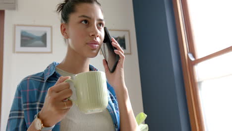 Happy-biracial-teenage-girl-drinking-tea,-talking-on-smartphone-at-home,-copy-space,-slow-motion