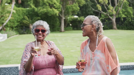 Glückliche-ältere-Afroamerikanische-Freundinnen-Mit-Sonnenbrille-Und-Getränken-Am-Pool-Sitzend,-Zeitlupe