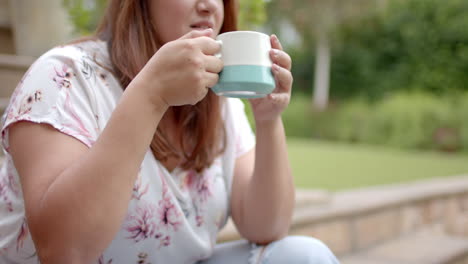 Happy-plus-size-biracial-woman-sitting-on-steps-in-garden-drinking-coffee,-copy-space,-slow-motion