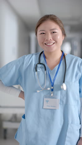 Vertical-video-of-portrait-of-happy-asian-female-doctor-in-hospital-corridor,-slow-motion