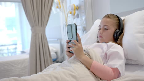 Caucasian-girl-patient-in-hospital-bed-in-headphones-using-smartphone,-copy-space,-slow-motion