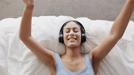 Happy-biracial-woman-lying-on-bed-using-headphones-and-listening-to-music,-slow-motion