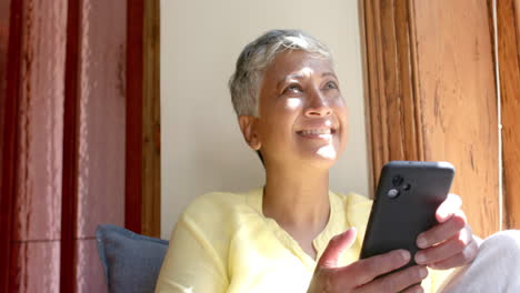 Happy-senior-biracial-woman-sitting-on-couch-and-using-smartphone-at-home,-slow-motion
