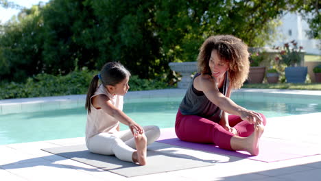 Happy-biracial-mother-and-daughter-practicing-yoga-sitting-stretching-in-sunny-garden,-slow-motion