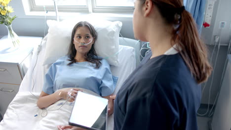 Diverse-female-patient-in-hospital-bed-and-female-doctor-using-tablet-talking,-slow-motion