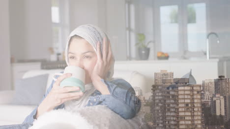 Animation-of-cityscape-over-biracial-woman-in-hijab-drinking-tea
