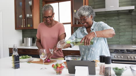 Happy-senior-african-american-female-friends-pouring-wine,-cooking-and-using-tablet,-slow-motion