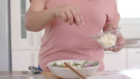 Happy-plus-size-biracial-woman-making-feta-cheese-and-vegetable-salad-in-kitchen,-slow-motion