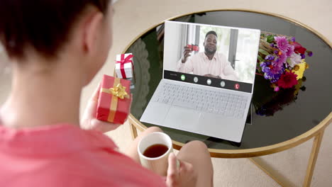 Caucasian-woman-holding-red-gift-using-laptop-with-african-american-man-on-screen