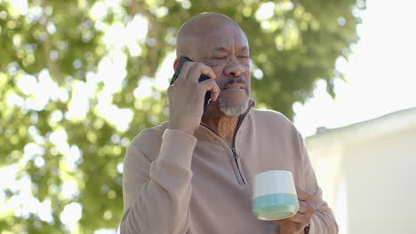Glücklicher-älterer-Mann-Mit-Gemischter-Abstammung-Auf-Dem-Balkon,-Der-Auf-Dem-Smartphone-Spricht-Und-Kaffee-Trinkt,-Zeitlupe