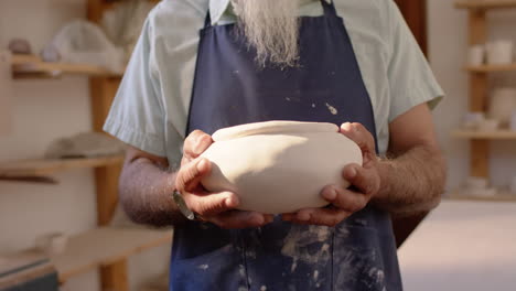 Mid-section-of-biracial-potter-with-long-beard-holding-bowl-in-pottery-studio,-slow-motion
