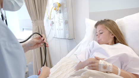 Caucasian-female-doctor-in-face-mask-using-stethoscope-on-girl-patient-in-hospital-bed,-slow-motion