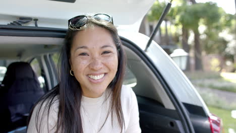 Glückliche-Asiatische-Frau-Mit-Sonnenbrille,-Die-Im-Kofferraum-Eines-Autos-Sitzt-Und-An-Einem-Sonnigen-Tag-Lächelt,-Zeitlupe