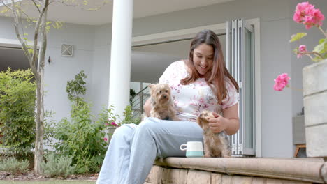 Happy-plus-size-biracial-woman-sitting-in-garden-with-coffee,-petting-dogs,-copy-space,-slow-motion