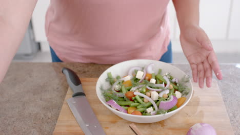 Happy-plus-size-biracial-woman-making-feta-cheese-and-vegetable-salad-in-kitchen,-slow-motion