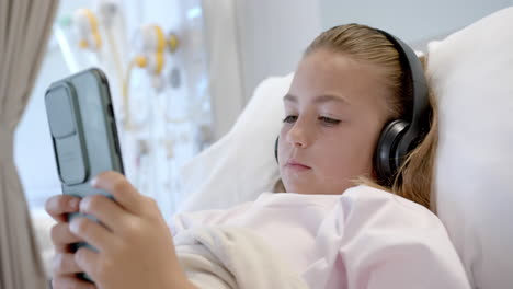 Caucasian-girl-patient-lying-in-hospital-bed-wearing-headphones-using-smartphone,-slow-motion