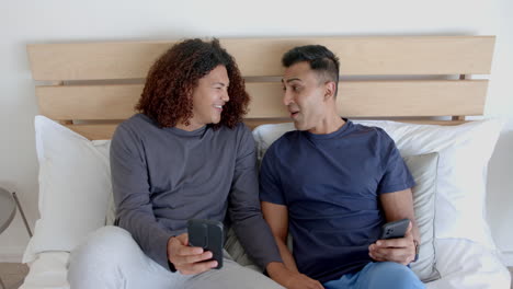 Happy-diverse-gay-male-couple-sitting-on-bed-using-smartphones,-talking-in-the-morning,-slow-motion