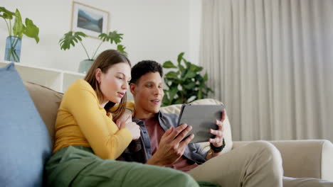 Happy-diverse-couple-sitting-on-sofa-using-tablet-at-home,-in-slow-motion