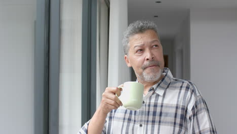 Thoughtful-senior-biracial-man-standing-at-window-drinking-coffee-at-home,-slow-motion