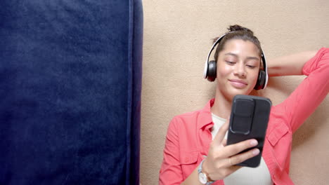 Smiling-biracial-teenage-girl-in-headphones-lying-on-floor-using-smartphone,-copy-space,-slow-motion