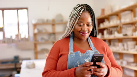 Happy-african-american-female-potter-using-smartphone-and-smiling-in-pottery-studio,-slow-motion