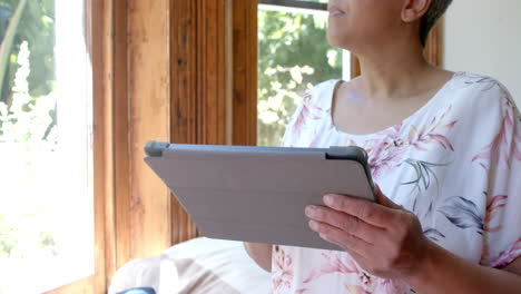 Nachdenkliche-ältere-Frau-Mit-Gemischter-Abstammung,-Die-Zu-Hause-Am-Fenster-Ein-Tablet-Benutzt,-Zeitlupe