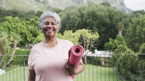 Portrait-of-happy-senior-african-american-woman-holding-yoga-mat-on-balcony,-copy-space,-slow-motion