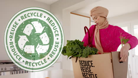 Biracial-woman-in-hijab-carrying-box-with-vegetables-in-kitchen-over-recycling-sign