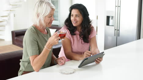 Happy-diverse-senior-women-using-tablet-and-drinking-tea-in-sunny-kitchen,-slow-motion