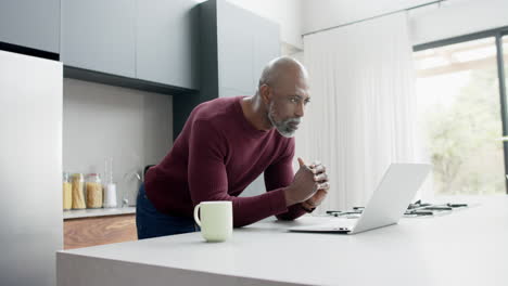 Happy-mature-biracial-man-with-cup-of-coffee-using-laptop-in-kitchen,-slow-motion