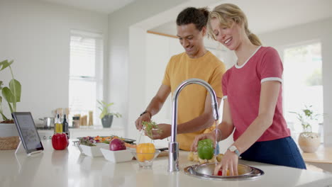 Happy-diverse-couple-preparing-and-washing-fresh-vegetables-in-kitchen-with-tablet,-slow-motion