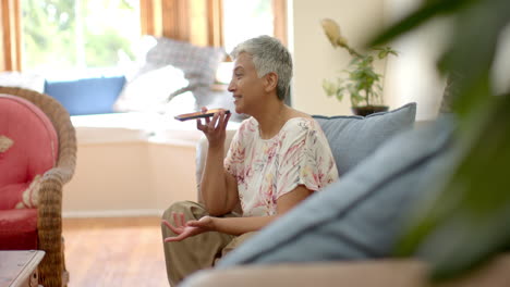 Glückliche-ältere-Frau-Mit-Gemischter-Abstammung,-Die-Auf-Der-Couch-Sitzt-Und-Zu-Hause-Mit-Dem-Smartphone-Spricht,-Zeitlupe