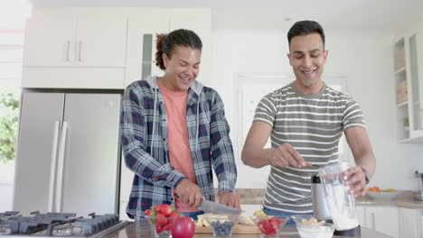Happy-diverse-gay-male-couple-preparing-healthy-fruit-smoothie-in-kitchen,-copy-space,-slow-motion