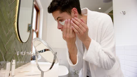 Focused-biracial-man-applying-under-eye-mask-in-bathroom-looking-in-mirror,-slow-motion