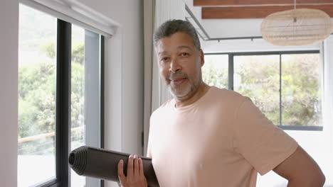 Portrait-of-happy-senior-biracial-man-holding-yoga-mat-at-home-and-smiling,-slow-motion