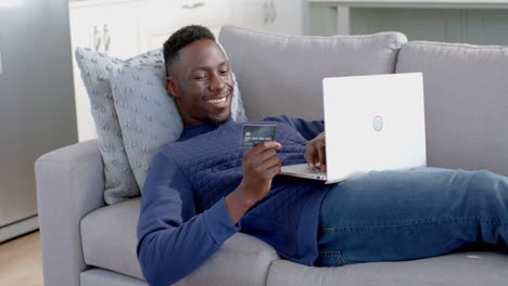 Glücklicher-Afroamerikanischer-Mann,-Der-Im-Sonnigen-Wohnzimmer-Liegt-Und-Laptop-Und-Kreditkarte-Benutzt,-Zeitlupe