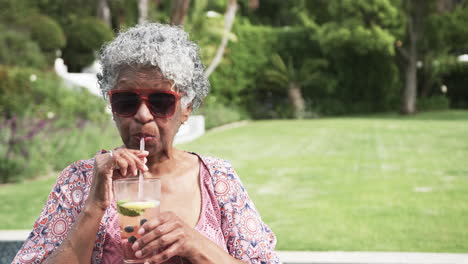Ältere-Afroamerikanische-Frau-Mit-Sonnenbrille-Trinkt-Cocktail-Im-Garten,-Kopierraum,-Zeitlupe